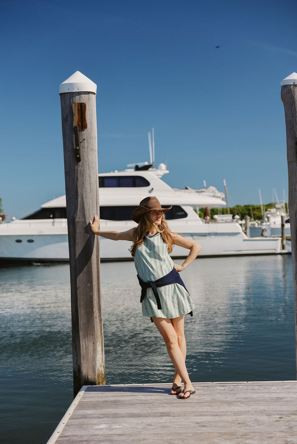 Mini Gili green stripe dress belted with navy sash at dock piling, Montauk Yacht Club marina and yacht in background, MTK Hamptons summer style 2026 Puka The Label
