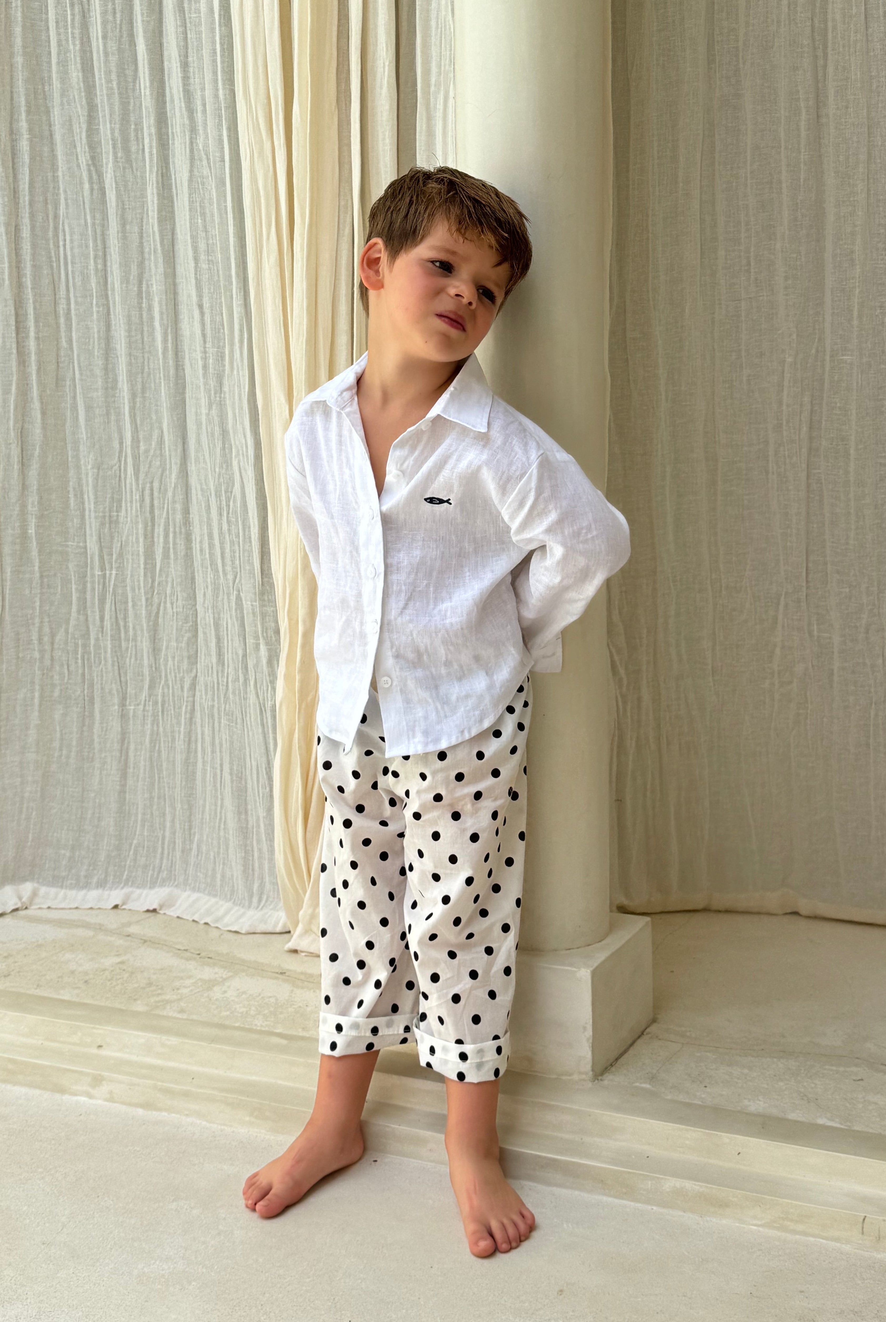 Boy wearing Pantai Polka Dot Pants with a white linen shirt, standing barefoot in a Bali resort villa — kids beach vacation outfit