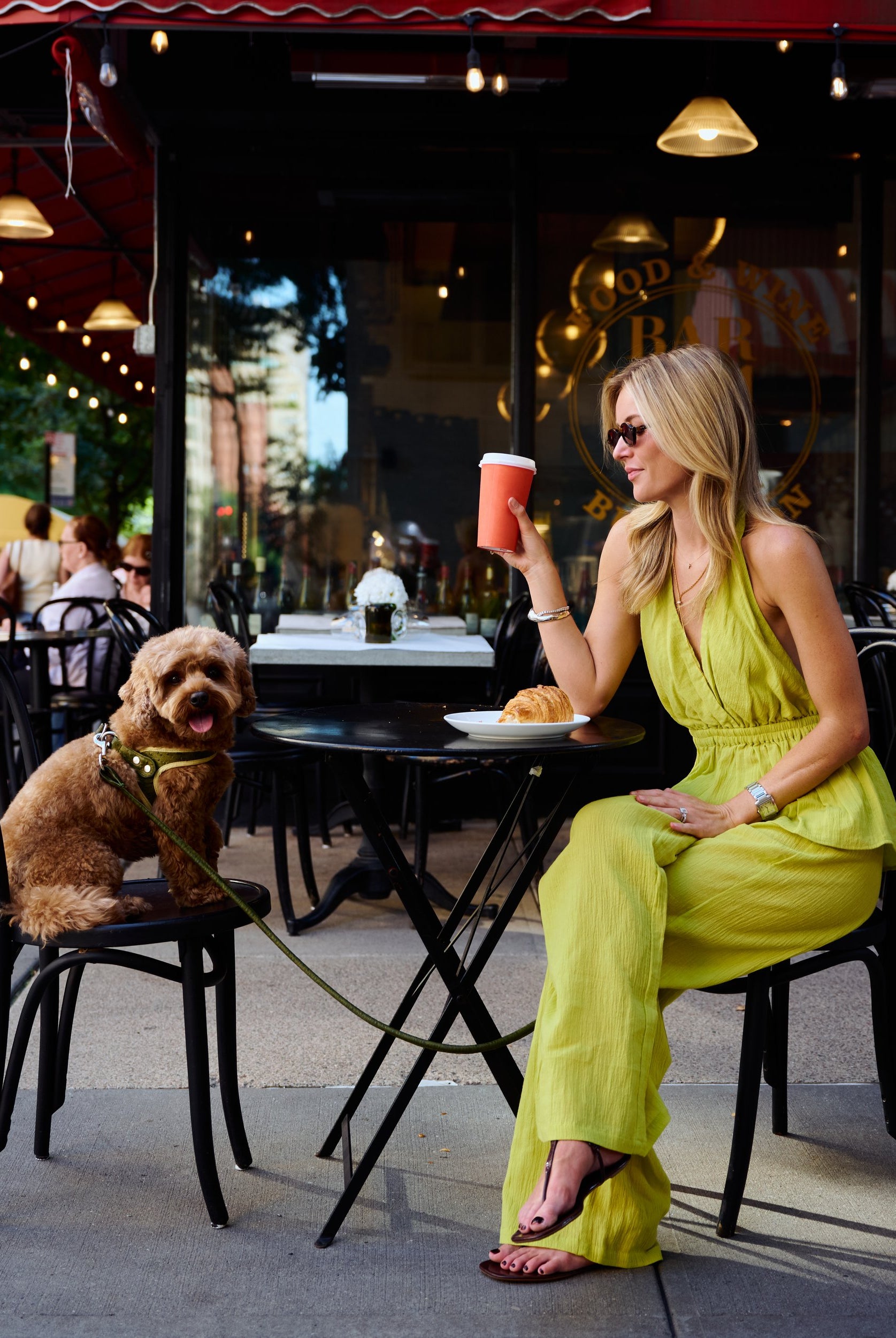 Natalie Simkins in Carlyle Set Olive Brine seated at NYC cafe holding coffee, curly dog on chair