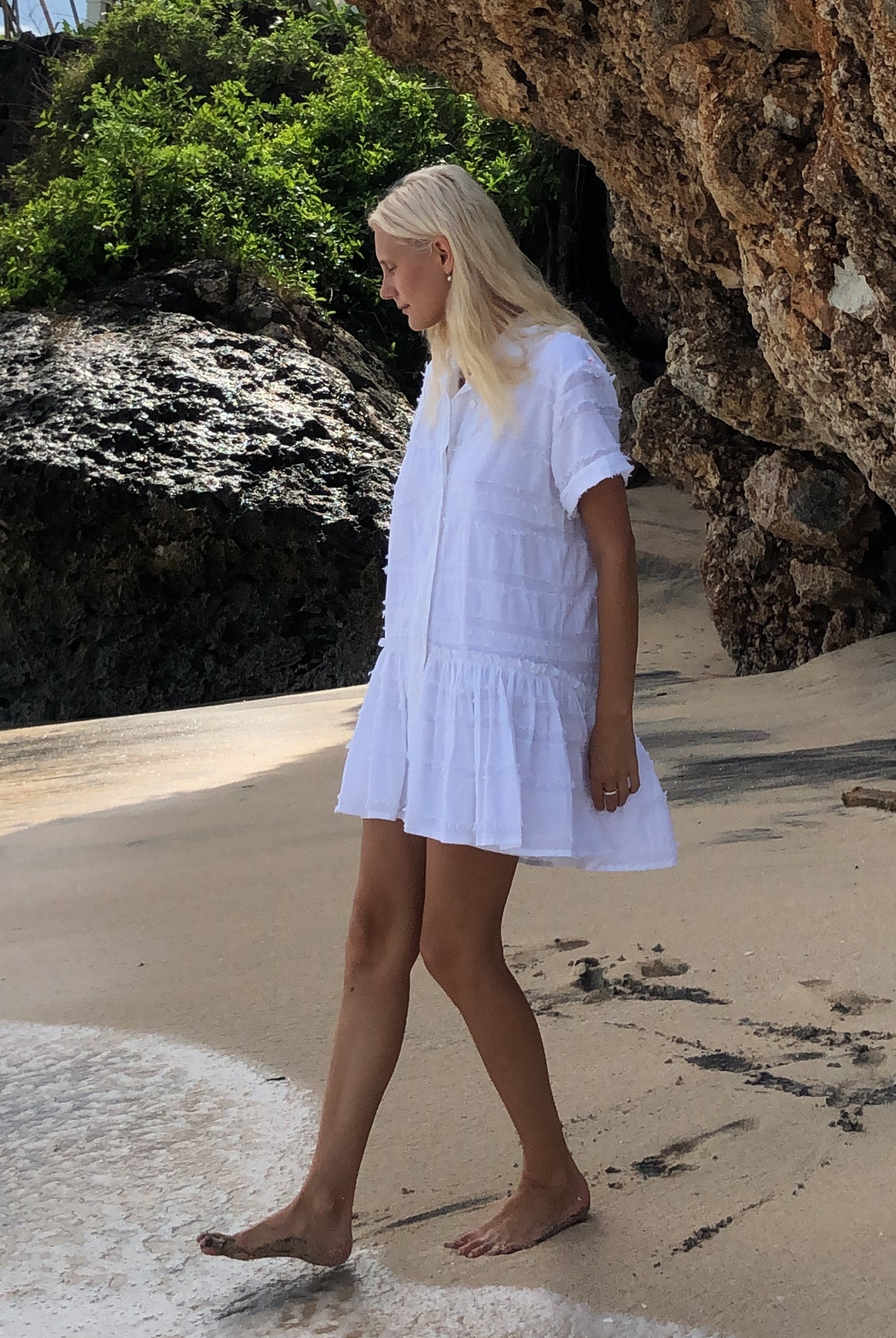 Person in a white dress standing on a rocky beach with ocean waves.