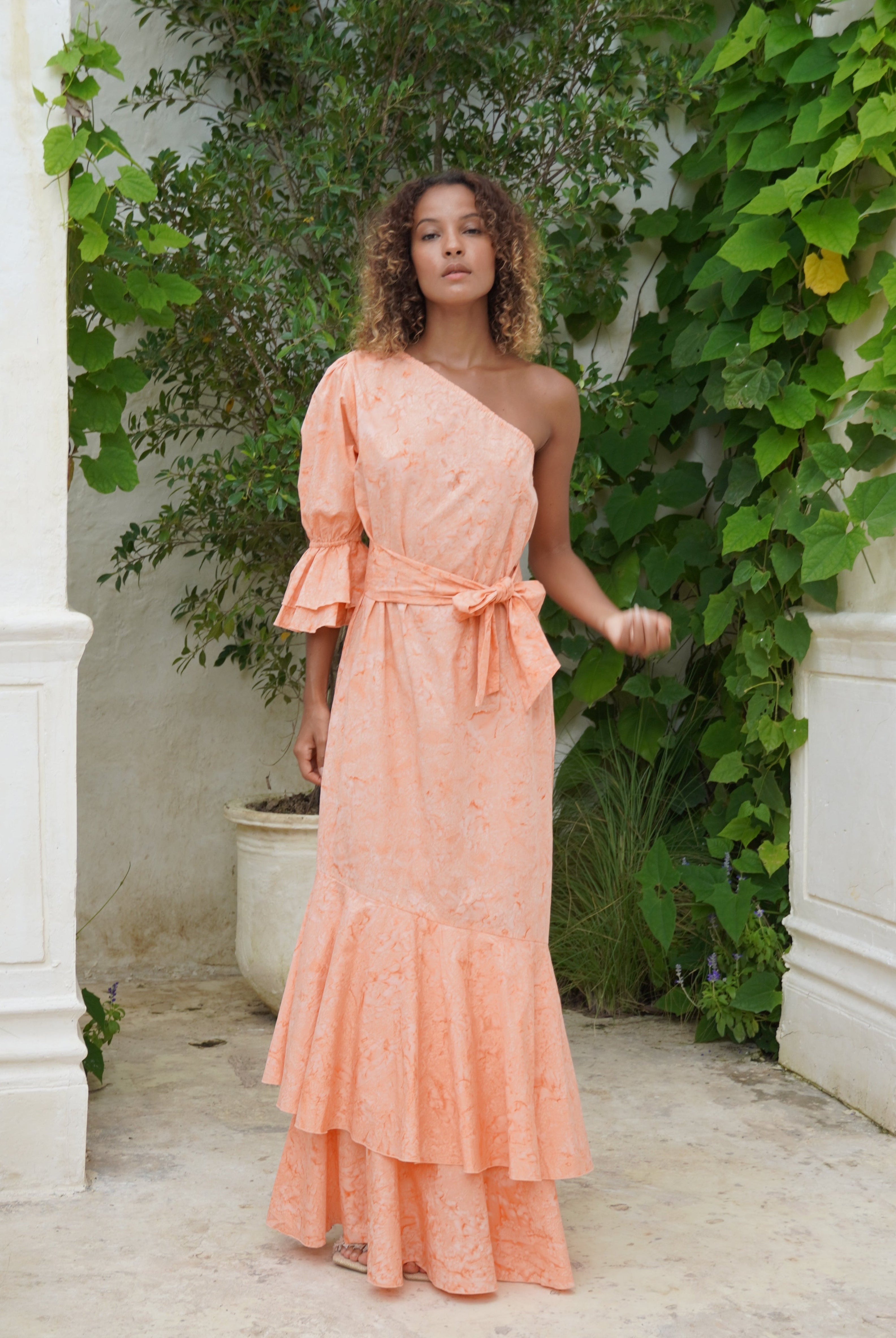 Woman in a peach ruffle dress standing in front of a white wall with greenery
