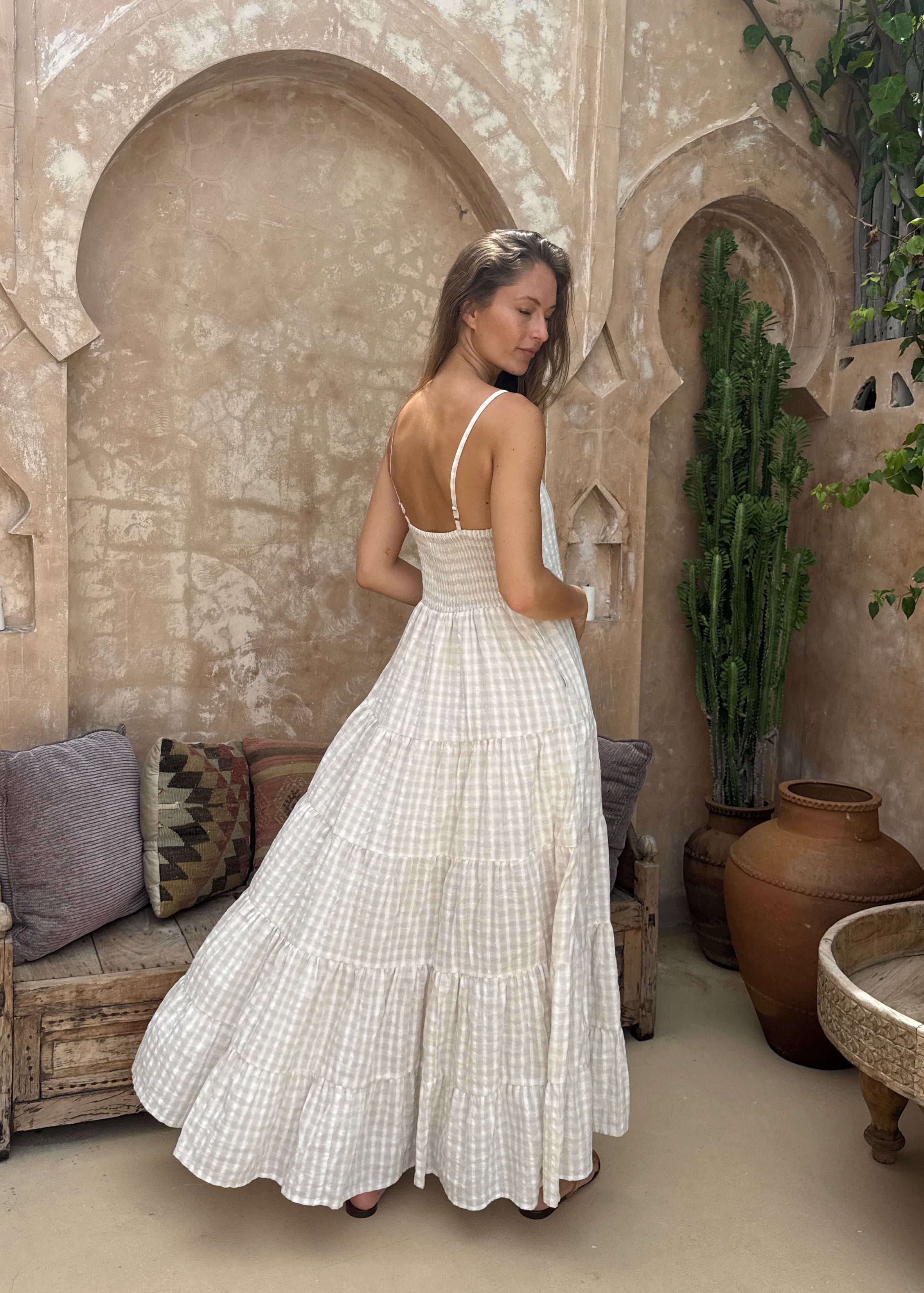 Woman in a white/beige dress standing in front of a stone archway with plants and a bench.