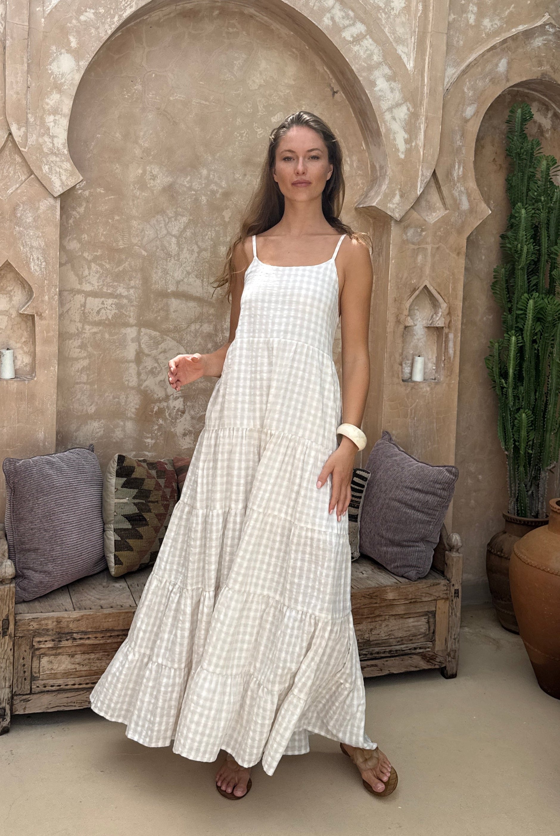 Woman in a beige dress standing in front of a decorative stone archway with plants and a wheel in the background.