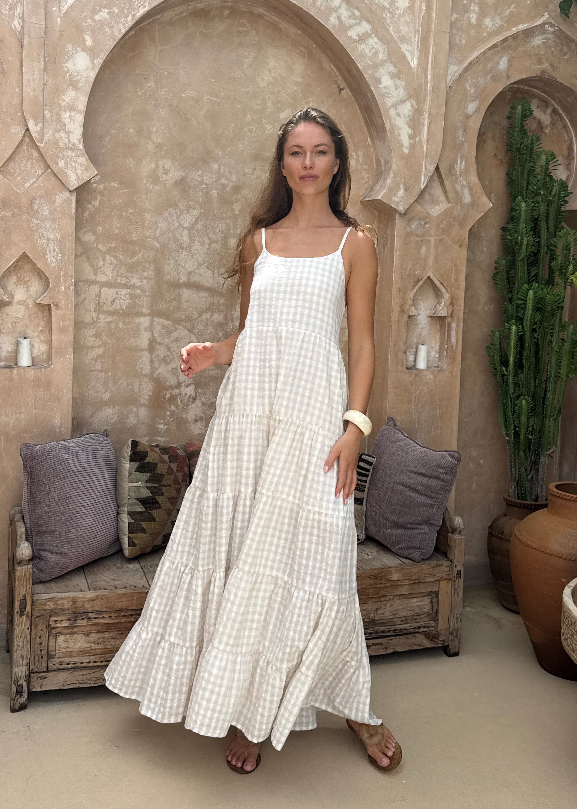 Woman in a beige dress standing in front of a decorative stone archway with plants and a wheel in the background.
