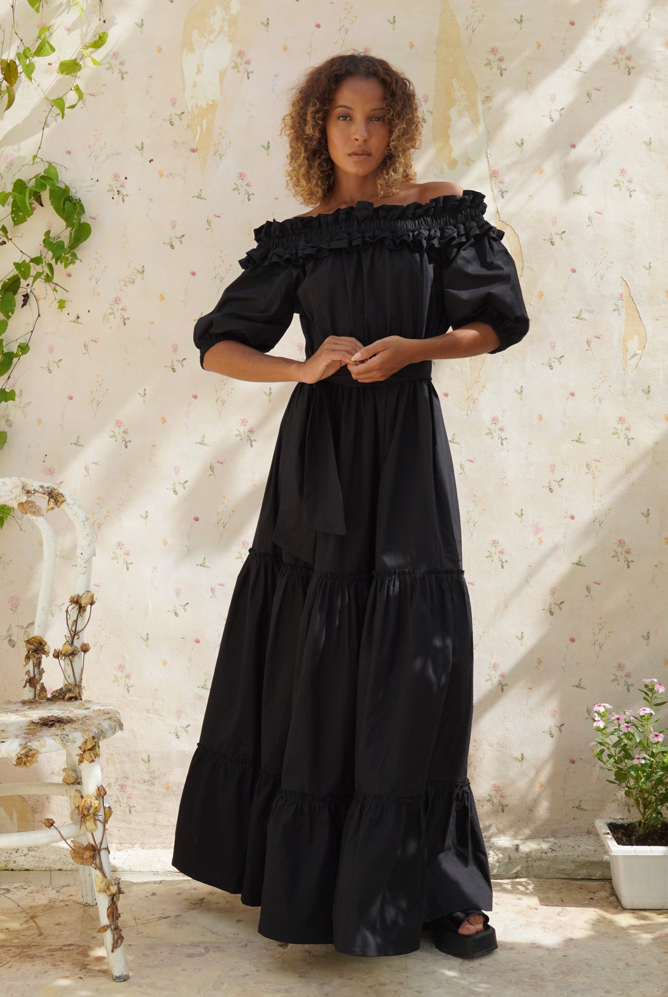 Woman in a black dress standing in a sunlit outdoor setting with white walls and greenery.