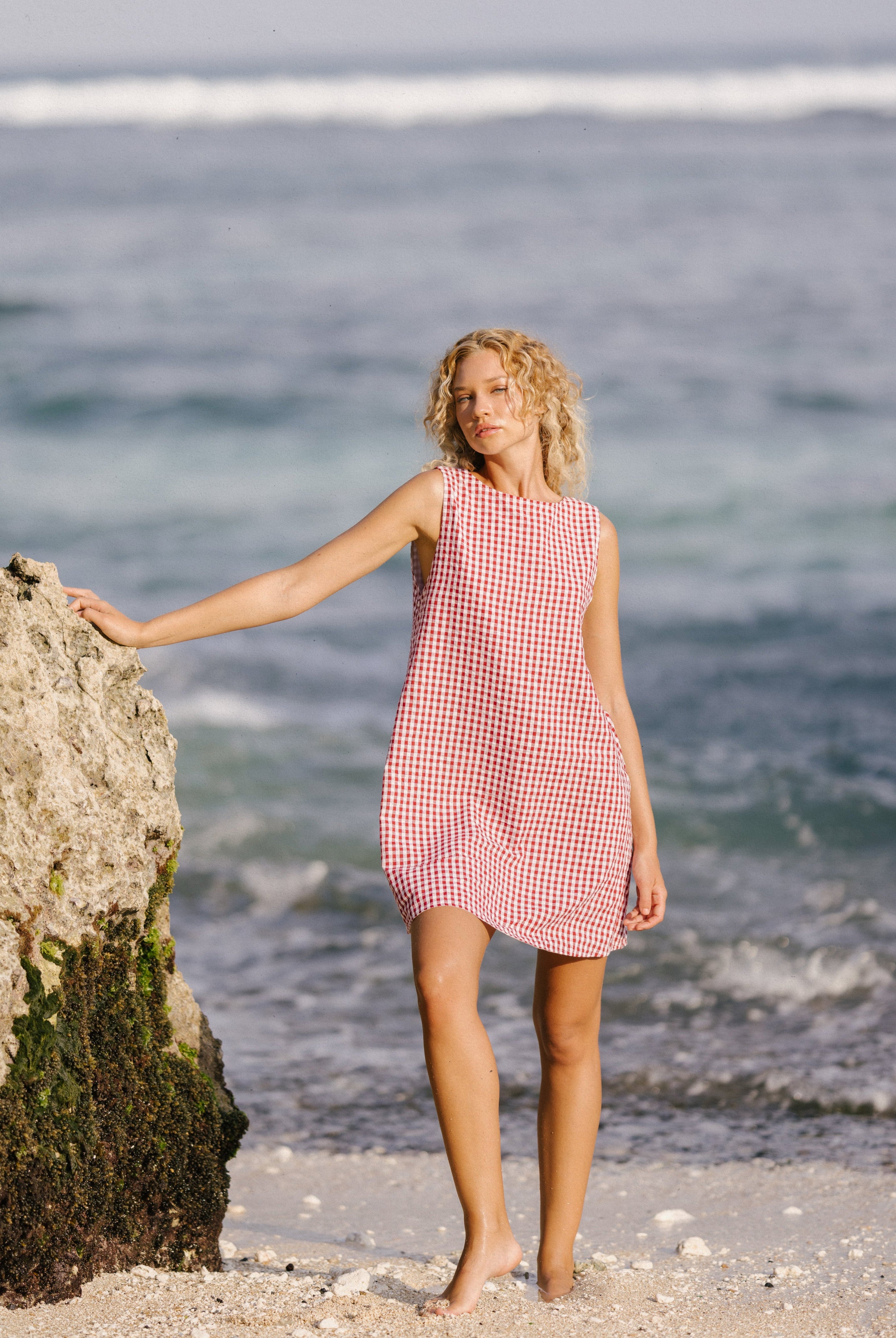 Woman in a red checkered dress standing on a beach with ocean waves in the background