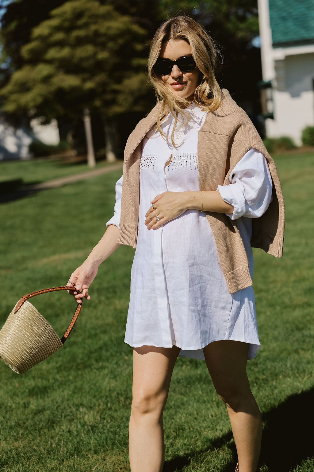 White linen Ulu Dress styled with camel knit cardigan and straw market bag on Montauk Yacht Club lawn, preppy MTK summer outfit idea 2026 Puka The Label