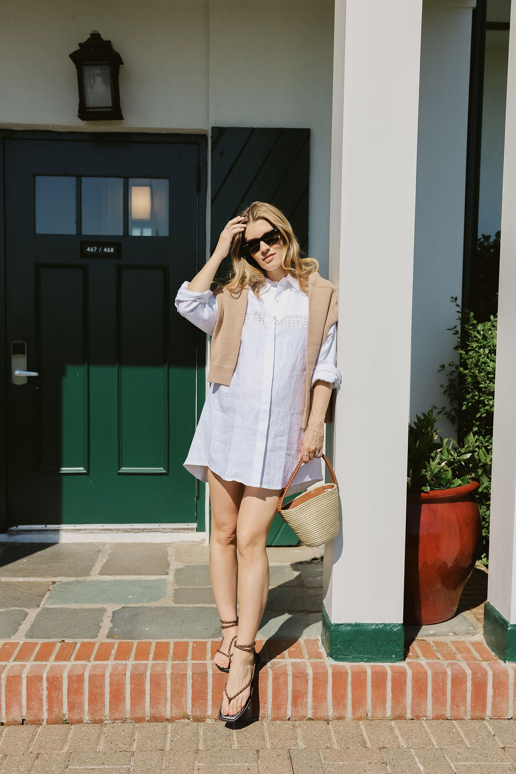 White linen Ulu Dress with black strappy sandals and straw basket bag in front of iconic Montauk Yacht Club green door, MTK summer outfit idea 2026