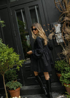 Jules Fox in a short dress, Fall NYC style inspiration, short black dress paired with stuart weitzman tall black boots. Black oversized ruffle sleeve mini dress NYC going-out outfit fall 2026, effortlessly cool cotton dress for city evening and downtown look