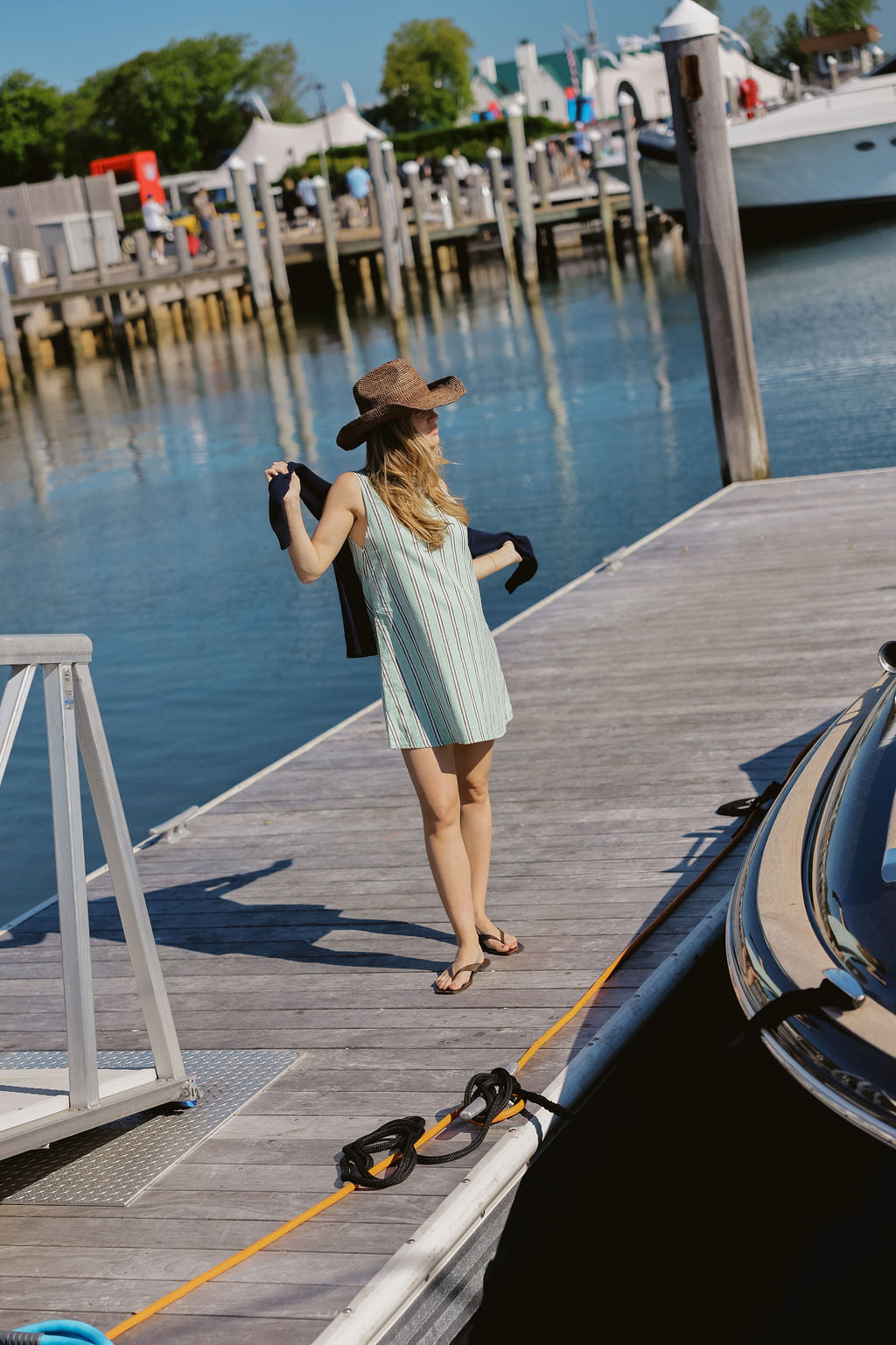 Back view of green stripe Mini Gili dress on Montauk Yacht Club marina dock, raffia hat and navy cardigan over shoulder, MTK nautical harbor summer outfit 2026