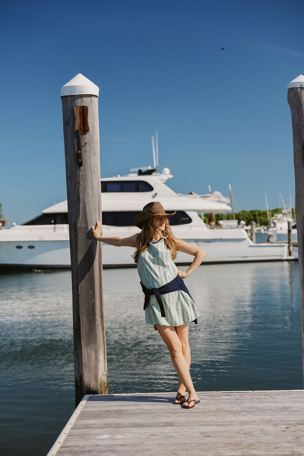 Mini Gili green stripe dress belted with navy sash at dock piling, Montauk Yacht Club marina and yacht in background, MTK Hamptons summer style 2026 Puka The Label