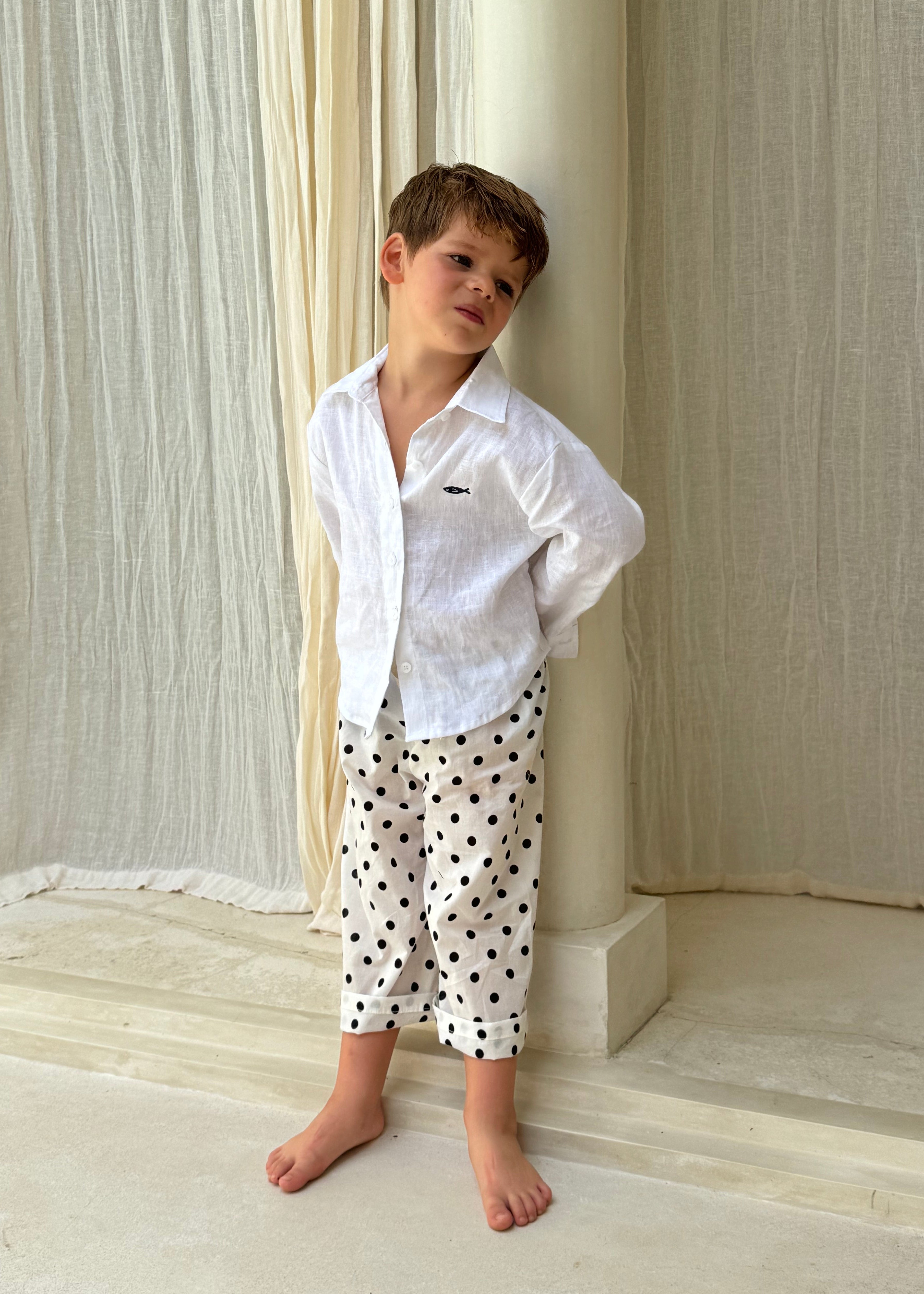 Boy wearing Pantai Polka Dot Pants with a white linen shirt, standing barefoot in a Bali resort villa — kids beach vacation outfit